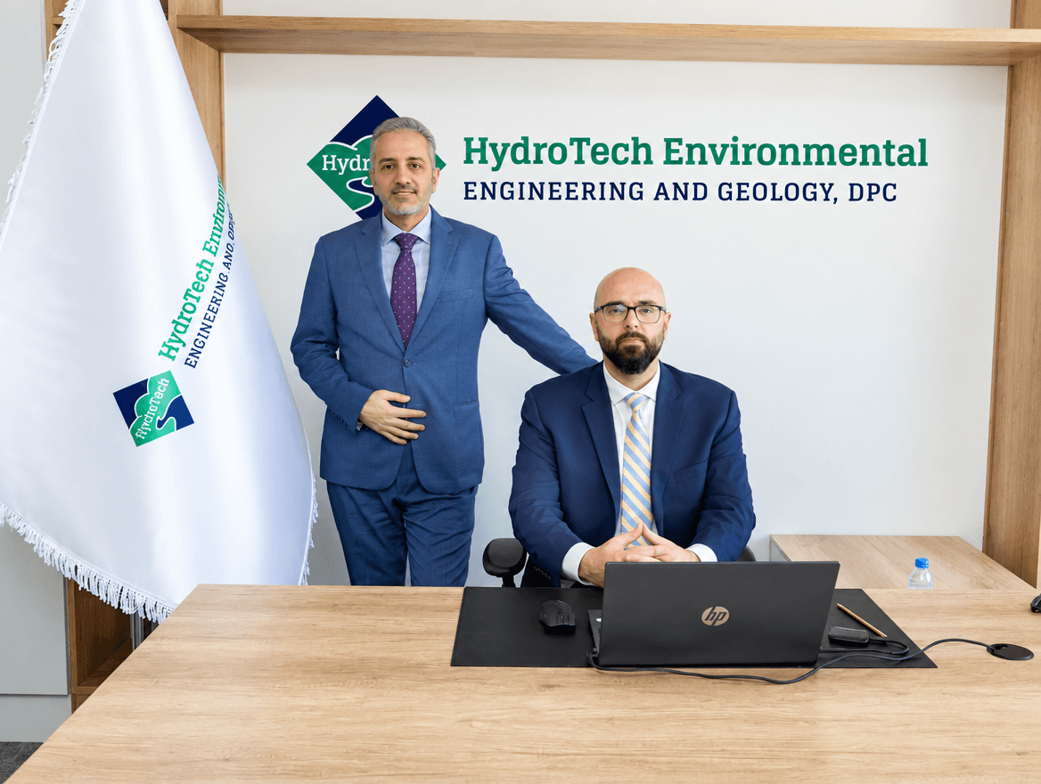 Two HydroTech Environmental Engineering and Geology executives in the new Riyadh office, standing beside the company flag and wall-mounted wordmark.