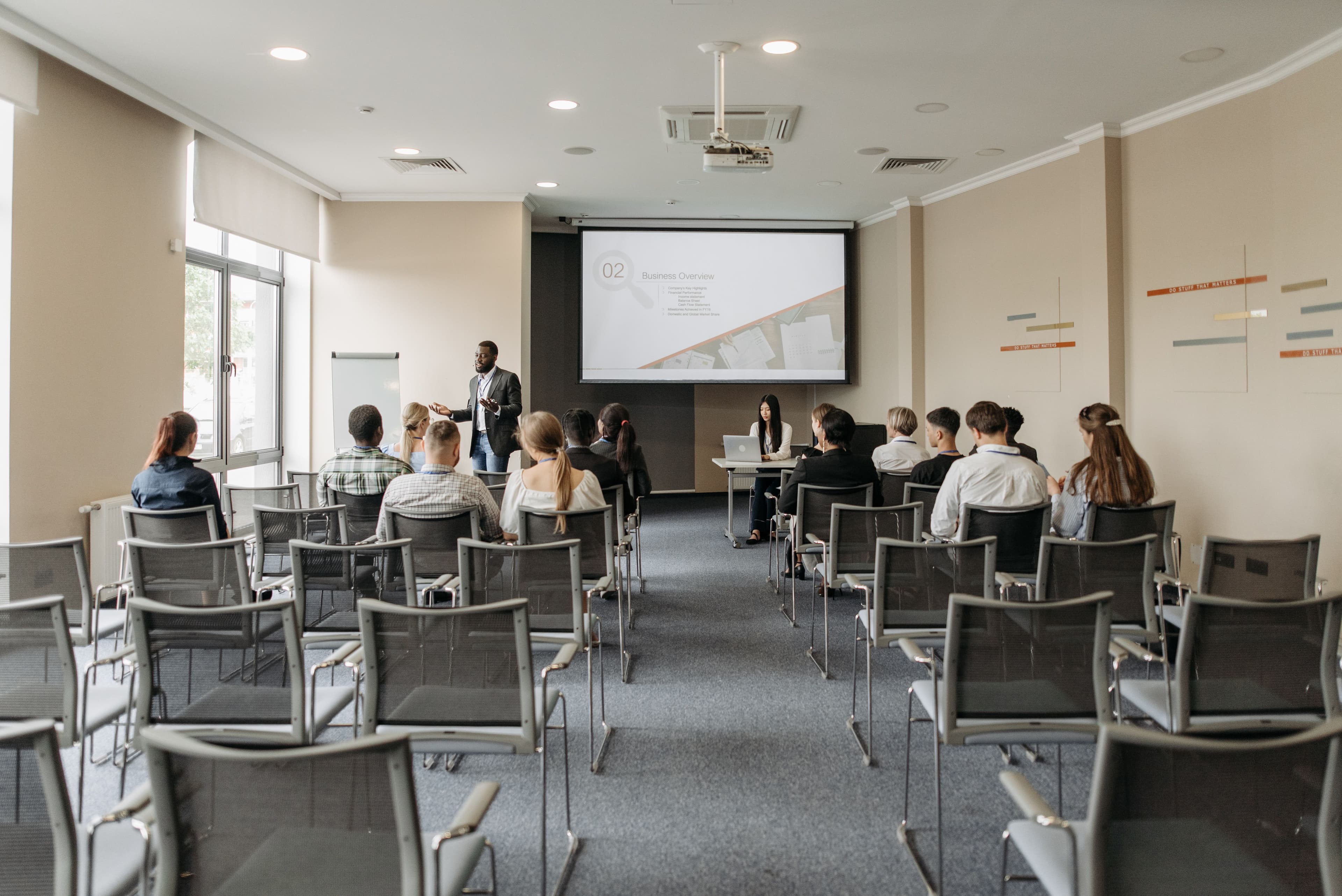 Trainer presenting to a seated group in a meeting room, with a slide projected on screen
