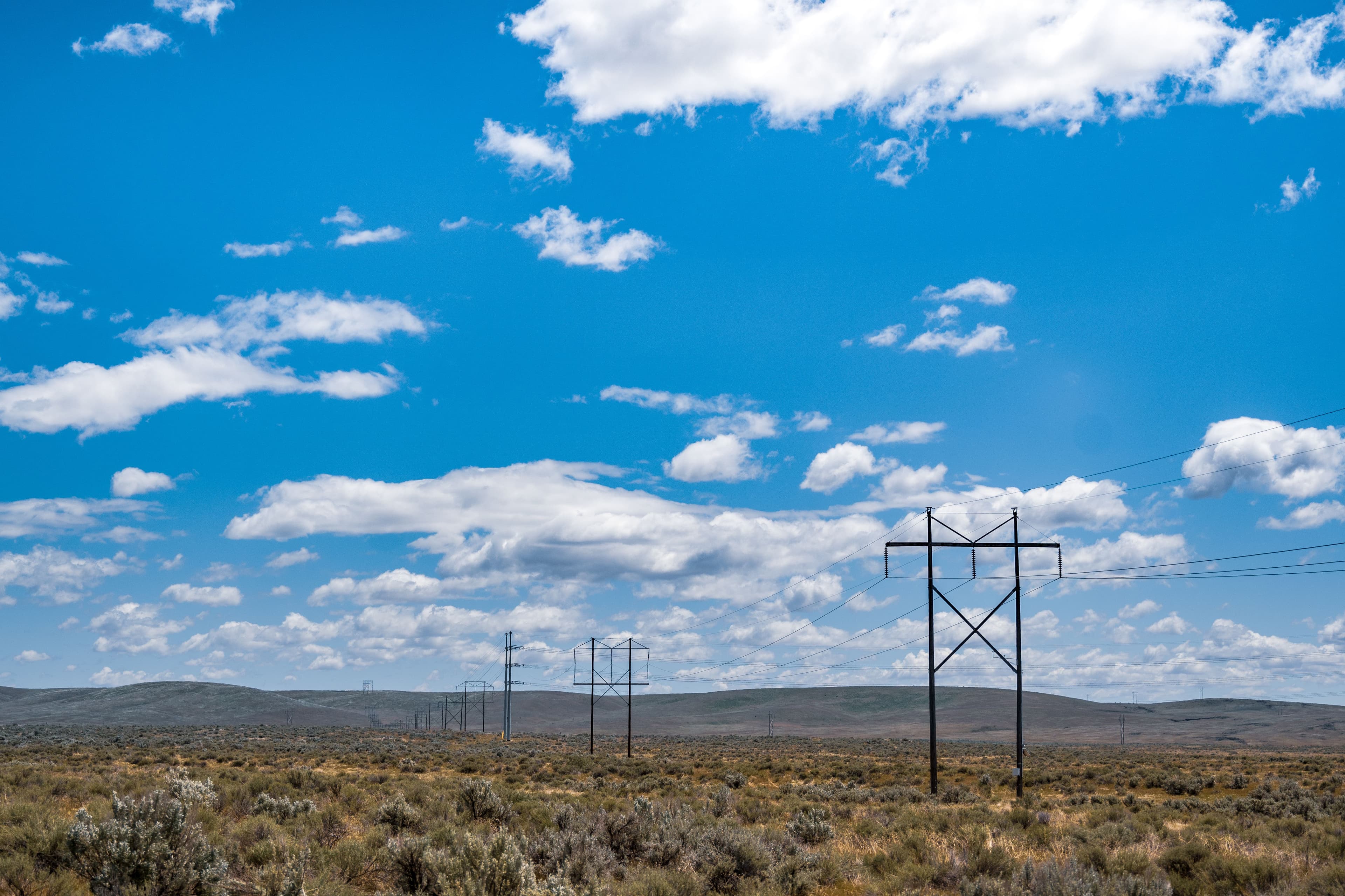 Row of steel transmission towers across dry scrubland under a blue sky with scattered clouds